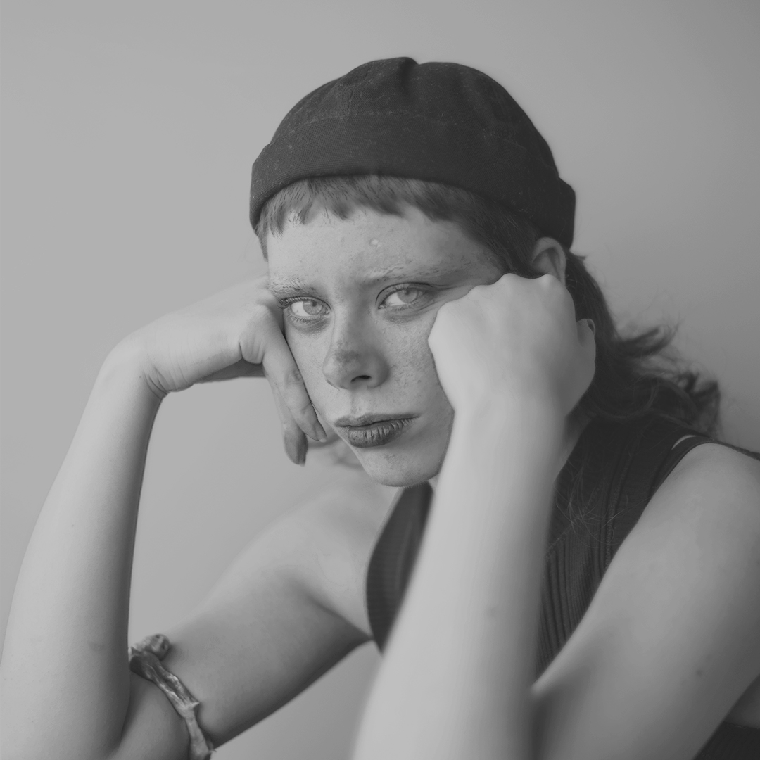 la foto contiene il ritratto di elisa Merra in bianco e nero. Ragazza giovene con le mani si mantiene la testa e fissa l'obiettivo. In testa ha un berretto nero dal quale esce una frangetta